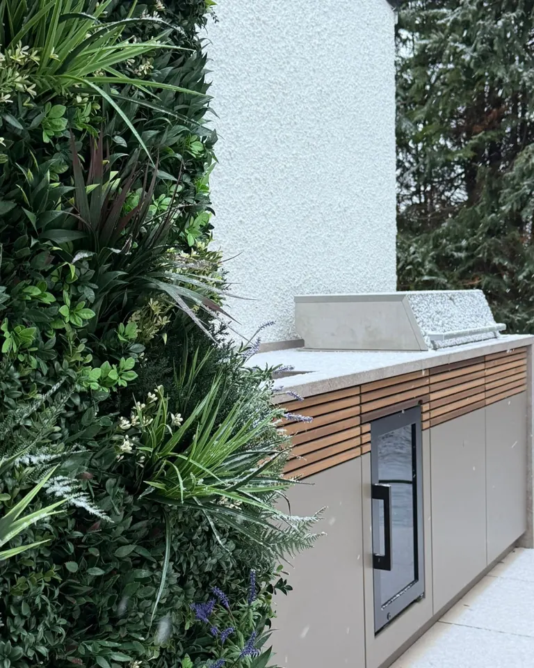 A Cubic outdoor kitchen with timber cladding and stone worktop beside a green wall, with snow falling