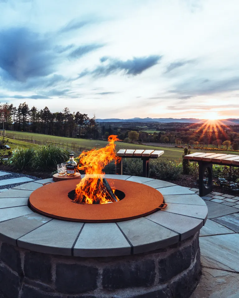 A corten fire pit set into a stone surround on a paved terrace, with open countryside and a setting sun beyond