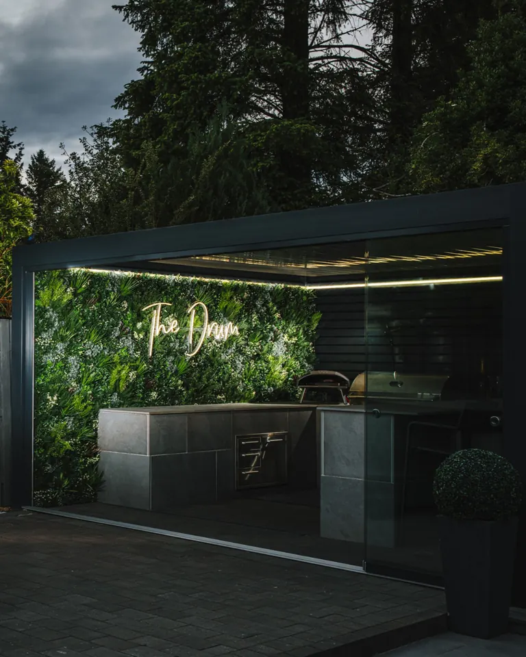 The Drum: a covered outdoor kitchen at dusk, with an illuminated sign on a green wall beneath a dark aluminium pergola